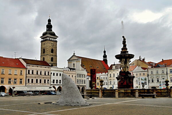 Marktplatz von Budweis mit steinigem Uhrturm, barockem Brunnen und moderner Skulptur vor bewölktem Himmel.