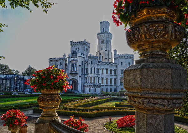 Historisches Schloss mit hohem Rundturm, formalem Garten und roten Blüten, im Vordergrund verzierte steinerne Blumengefäße.