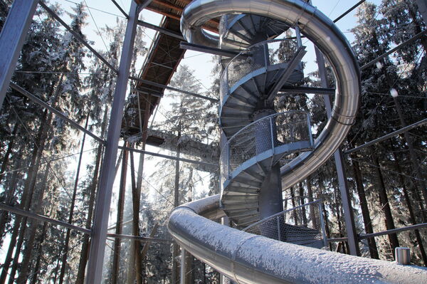 Metallspiraltreppe mit großer Edelstahlrutsche an einem erhöhten Baumwipfelpfad, verschneite Tannen im Wald