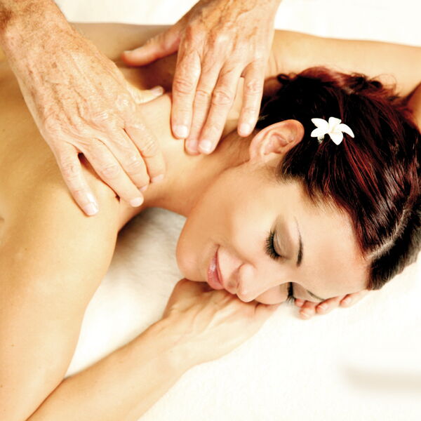 A woman is lying relaxed on her stomach and receiving a neck and shoulder massage from Kahraman. She has a white flower in her hair.